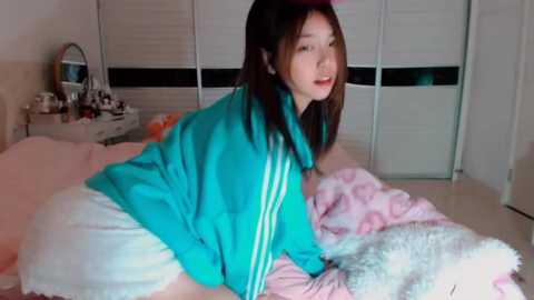 Media: Video of an East Asian woman in a teal jacket and white skirt, lying on a bed with pink and white blankets. Background features a white wardrobe and a vanity mirror.