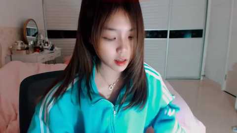 Media: Video of an Asian woman with straight black hair, wearing a turquoise tracksuit, sitting in a bedroom with white walls, a vanity table, and a closed wardrobe.