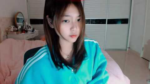 Media: Video of a young Asian woman with straight, shoulder-length brown hair, wearing a turquoise jacket and sitting on a bed with white walls and a closed window.