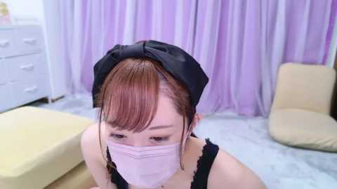 Media: Video of a young East Asian woman in a black headscarf, wearing a pink surgical mask, standing in a pastel-colored room with light purple curtains, white dresser, and beige furniture.