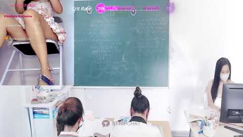 Media: Video of an office setting with two women in casual attire facing a green chalkboard displaying text. One woman sits, legs crossed, while the other stands at a desk, both wearing masks.