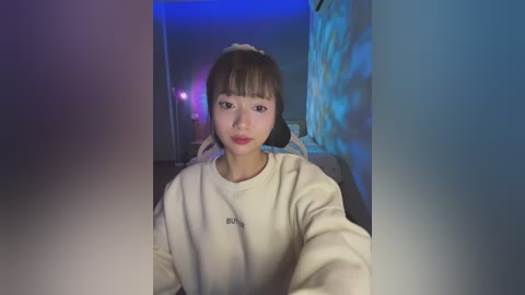 Media: Video of an East Asian woman with straight black hair, fair skin, and a neutral expression, wearing a beige sweater. Background features a dimly lit room with blue and purple lighting and a textured wall.