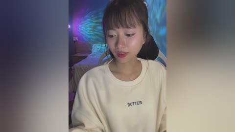 Media: A video of an Asian woman with straight brown hair and light skin, wearing a beige \"Butter\" sweatshirt, standing indoors with a colorful LED wall behind her.