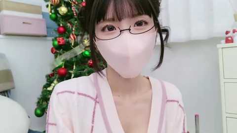 Media: A video of an Asian woman with straight black hair, wearing a pink face mask, round glasses, and a light pink robe, standing in front of a decorated Christmas tree with red and gold ornaments.