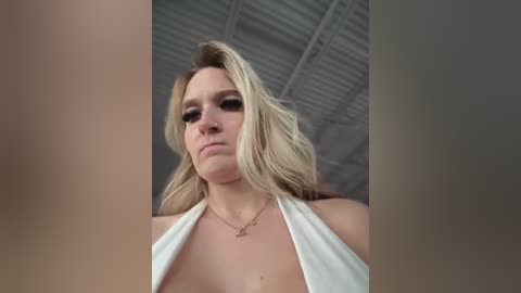 Media: Video of a blonde woman with a confident expression, wearing a white halter top, standing in an industrial setting with a metallic ceiling.