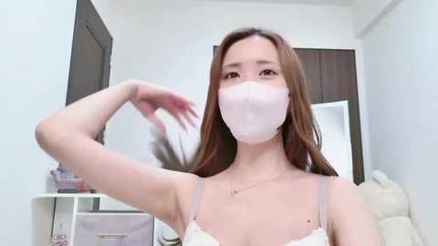 Media: A video of an Asian woman with long brown hair, wearing a white face mask, white lace bra, and a gold necklace, posing in a modern, white-walled bedroom.