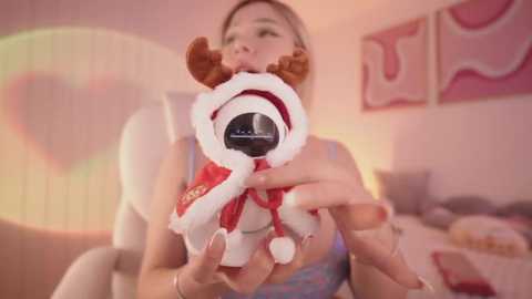 Media: Video of a blonde woman wearing a red Santa hat, holding a plush toy dog with a black face. The background features soft pink hues and abstract wall art.