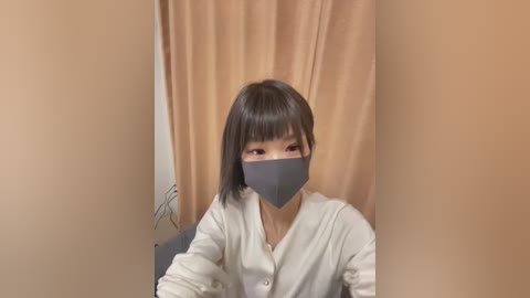 Media: A video of an Asian woman with shoulder-length black hair, wearing a gray face mask and white cardigan, sitting in a beige-curtained room.