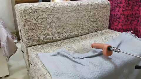 Media: Video of a beige couch with a textured pattern, covered in a light blue blanket. A sander with a wooden handle is applying a light gray paint to the fabric, revealing the texture beneath. The background includes a red curtain and a partially visible person in the distance.