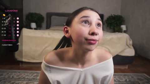 Media: A video of a young Asian woman with long black hair, wearing a white off-shoulder top, standing in a modern bedroom with a gray headboard and potted plants.