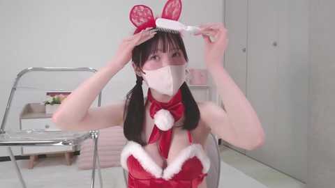 Media: Video of an Asian woman with long black hair in a red bunny outfit with white fur trim, adjusting a red headband. She wears a white mask, standing in a minimalist, white-tiled room.