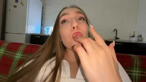 Media: Video of a young woman with long, straight, light brown hair, fair skin, and green eyes, wearing a white shirt, licking her finger, in a modern kitchen with a red plaid couch in the background.