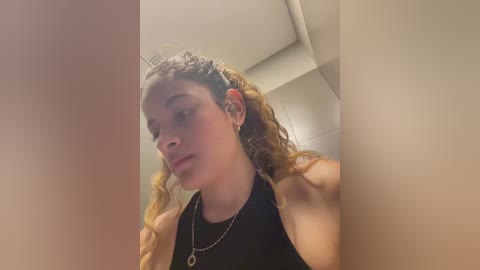 Media: A video of a young woman with light skin and curly, shoulder-length brown hair, wearing a black halter top and gold necklace, taken indoors with a blurry, beige wall background.