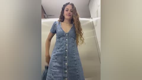 Media: Video of a young woman with curly, wavy brown hair, wearing a light blue, short-sleeved denim dress with buttons, standing in a narrow, dimly lit hallway.