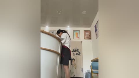 Media: Video of a young woman with long black hair, wearing a white blouse and black skirt, bending over a sink in a modern, minimalist bathroom with beige walls and framed artwork.