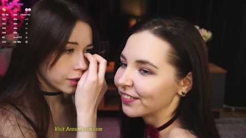 Media: Video of two young women with light skin, one applying makeup to the other's eye. They have dark hair, wearing black choker necklaces, in a dimly lit room.