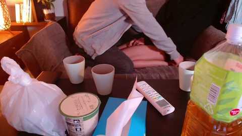 Media: Video of a cozy living room with a person in a gray sweater, drinking from a white cup. Nearby, there's a white plastic bag, a can of orange juice, a remote control, and a blue piece of paper on a dark wooden table.