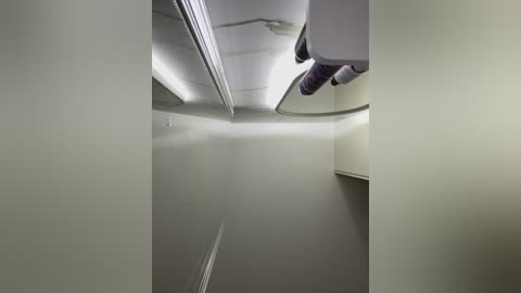 Media: Video of a narrow, empty, white-tiled corridor with a sloped ceiling, featuring a fluorescent light strip, a white pipe, and a purple hose hanging from the ceiling.