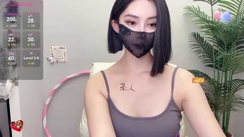 Media: Video of a young Asian woman with straight black hair, wearing a gray tank top and black face mask. She sits indoors with a hula hoop and plant in the background.