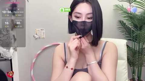 Media: Video of an Asian woman with shoulder-length black hair, wearing a black mask, adjusting a pink and white balloon bracelet in a room with a white couch, a green plant, and a framed picture.