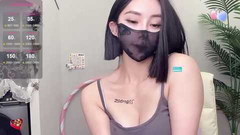 Media: Video of an East Asian woman with straight black hair, wearing a grey tank top and a black face mask, showing tattoos on her collarbone. Background includes a plant and a wall-mounted clock.