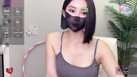 Media: Video of an Asian woman with shoulder-length black hair, wearing a gray tank top, black face mask, and gold hoop earrings, indoors, with a green plant and a hula hoop in the background.