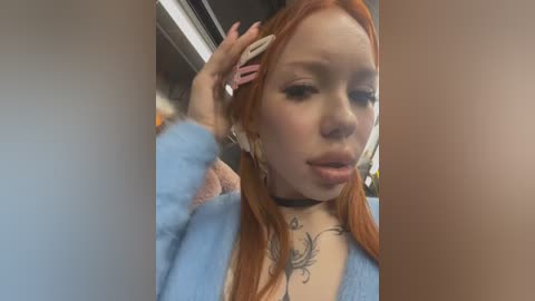 Media: A close-up video of a young woman with long, straight red hair, wearing a light blue cardigan, a black choker, and white headphones. She has a tattoo on her chest and is touching her hair.