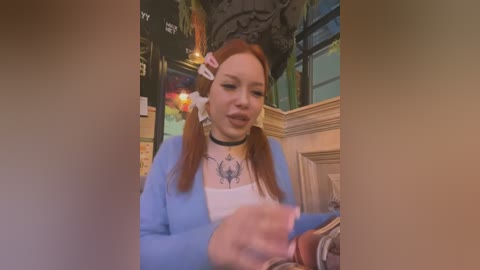 Media: Video of a young woman with straight, light brown hair styled in pigtails, wearing a light blue cardigan over a white shirt with a black tattoo sleeve design. She is seated in a cozy, dimly lit caf\u00e9 with a mix of wooden and green accents.