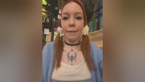 Media: Video of a pale-skinned woman with red pigtails, wearing a white top and blue cardigan, with a black choker featuring intricate floral tattoo designs on her chest, inside a cozy, dimly lit cafe.