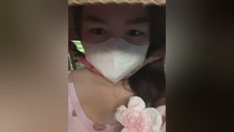 Media: A video of an Asian woman with medium skin tone wearing a white face mask, holding a pink flower, taken indoors with a blurred background.