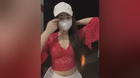 Media: A video of an Asian woman with long black hair, wearing a white cap, red lace top, and white pants, posing indoors with a red background, holding a mask.