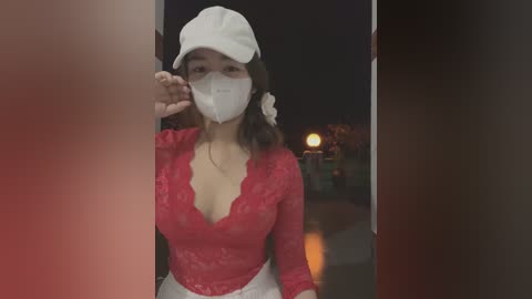 Media: Video of a young Asian woman with medium skin tone and long dark hair, wearing a white surgical mask, red lace top, white headband, and white pants, standing in a dimly lit doorway.