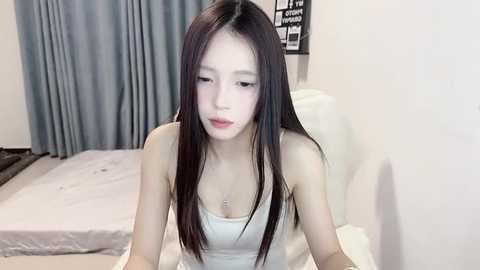 Media: Video of an Asian woman with long black hair, fair skin, and small breasts, wearing a white tank top, sitting on a bed with white sheets, in a bedroom with blue curtains.