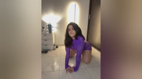 Media: Video of a Latina woman with long dark hair, wearing a purple mesh long-sleeve top and shorts, kneeling on a tiled floor, surrounded by a dimly lit bedroom with a nightstand and lamp.