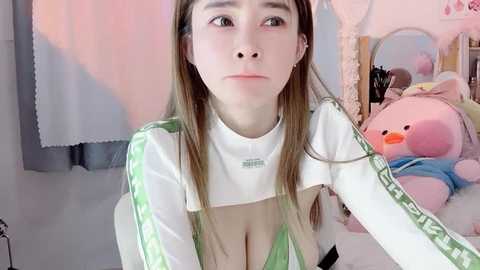 Media: Video of an East Asian woman with long brown hair, fair skin, and a slender build, wearing a revealing green-and-white sports top, in a pink-themed room with plush toys.