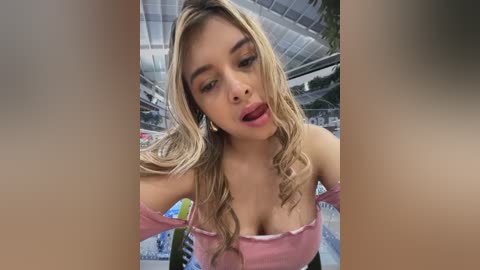 Video of a young woman with long, wavy blonde hair, fair skin, and full lips, wearing a pink off-shoulder top, taken in a swimming pool area.