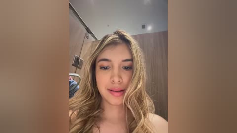 Media: Video of a young woman with long, wavy blonde hair and light skin, wearing minimal makeup, in a bathroom with wooden walls and a mirror.