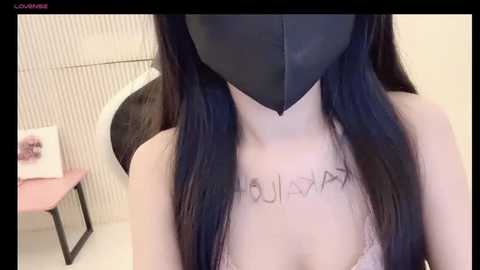 Media: Video of a person with long black hair wearing a black mask, revealing the word \"HOULIYAN\" tattooed on their chest, standing in a minimalist room with a white wall and a chair.