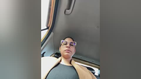 Video of a woman in glasses, wearing a gray sweater and beige jacket, reclining in a car seat with a beige headrest, partially visible through a window.