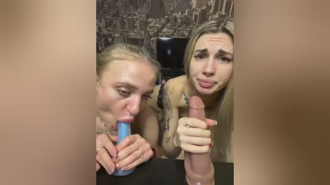 Media: Video of two women, one with blonde hair, the other with long brown hair, both holding and sucking on large, erect penises. Background features a dark, abstract mural.