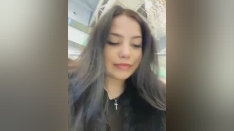 Video of a young woman with long, dark hair, wearing a black jacket and a cross necklace, standing indoors with blurred background.