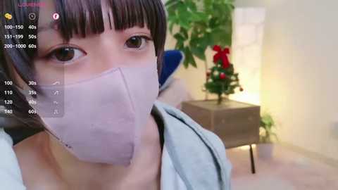 Media: A video of an East Asian woman with straight black hair, wearing a surgical mask, light blue top, and a red Christmas tree in the background.