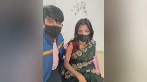 Media: Video of a young man in a blue and yellow shirt with a pink tie, and a woman in a green sari with a black mask, sitting indoors, with a child's drawing on the wall.