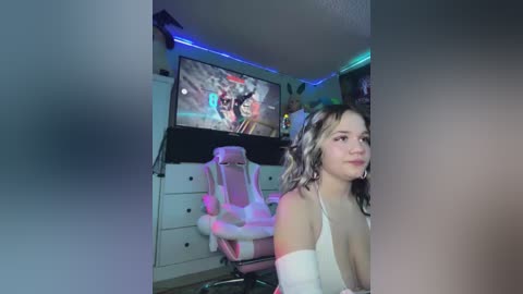 Media: A video of a young woman with wavy blonde hair, wearing a white top, sitting in a gaming chair in a dimly lit room with neon lights, TV showing game footage.