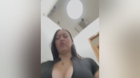 Media: A video of a woman with light skin, dark hair, and a low-cut black top, captured from a low angle, revealing her cleavage. The background features a white ceiling with a circular light fixture and a wooden door.