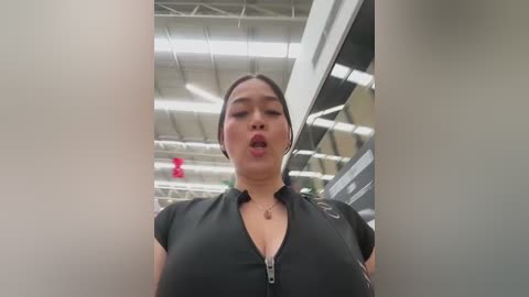 Media: Video of an Asian woman with medium skin tone, dark hair pulled back, wearing a black shirt with a plunging neckline, standing in a modern, brightly lit indoor space.