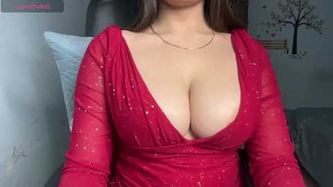 Media: A video of a woman with light skin, long brown hair, wearing a revealing, red, sequined dress that reveals her ample cleavage. The background features a gray couch and decorative wall art.