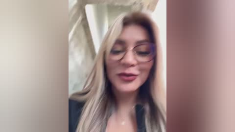 Media: Video of a fair-skinned woman with long blonde hair, wearing glasses, looking downward. She has a slender physique and is dressed in a dark top. The background is blurred, with a hint of a beige curtain.