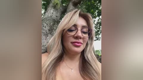 Media: Video of a young woman with light skin, long blonde hair, and round glasses, wearing a pink necklace, standing outdoors near a tree.