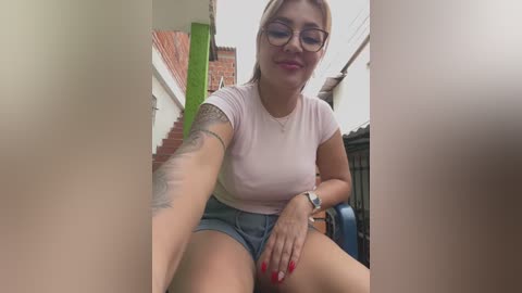 Media: Video of a woman with light skin, glasses, and pink top, sitting on a chair, showing a tattooed arm, against a narrow, brightly lit alleyway with red brick buildings.